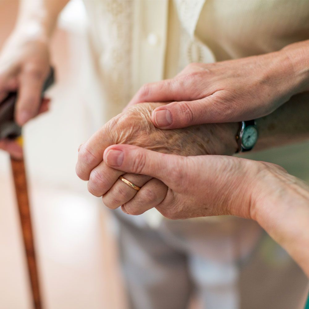 Handhalten einer älteren Person, die einen goldenen Ring trägt, im Hintergrund ist ein Gehstock zu sehen
